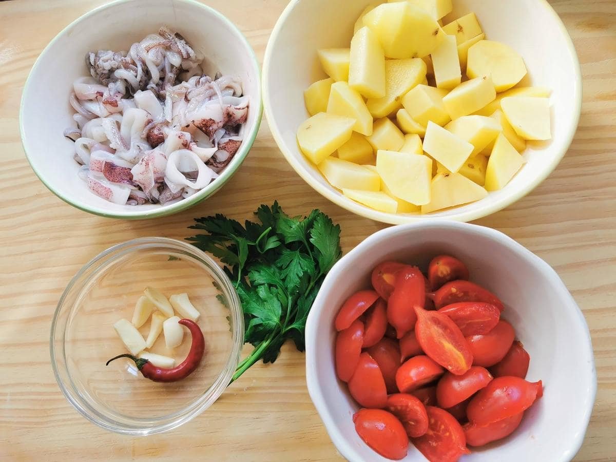 Prepared calamari, garlic, chilli potatoes and cherry tomatoes in bowls. Flat parsley on a wooden board.