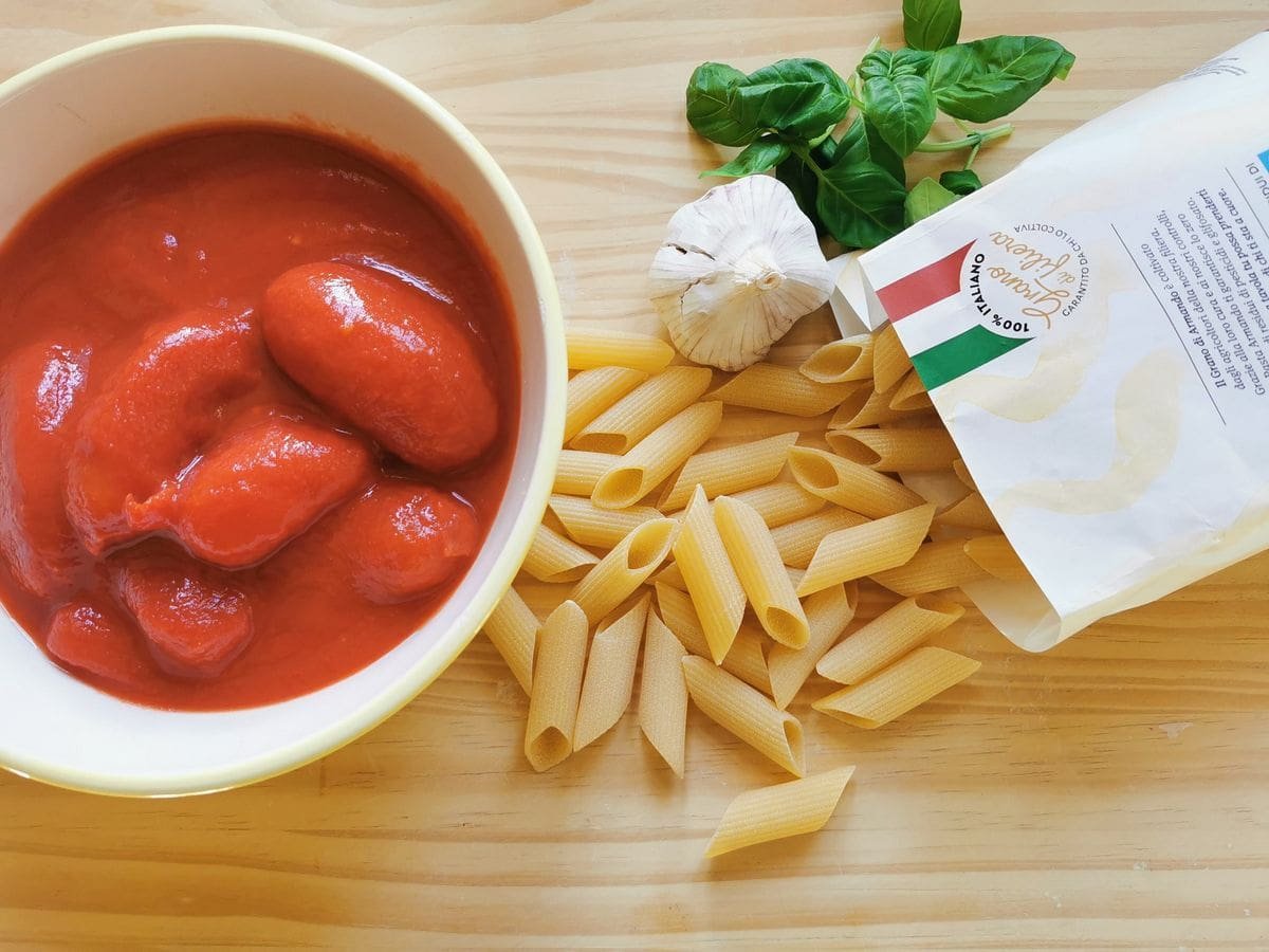 Ingredients for Penne Pomodoro on woodworking surface; Can of tomatoes, pens pasta, fresh basil leaves and garlic.