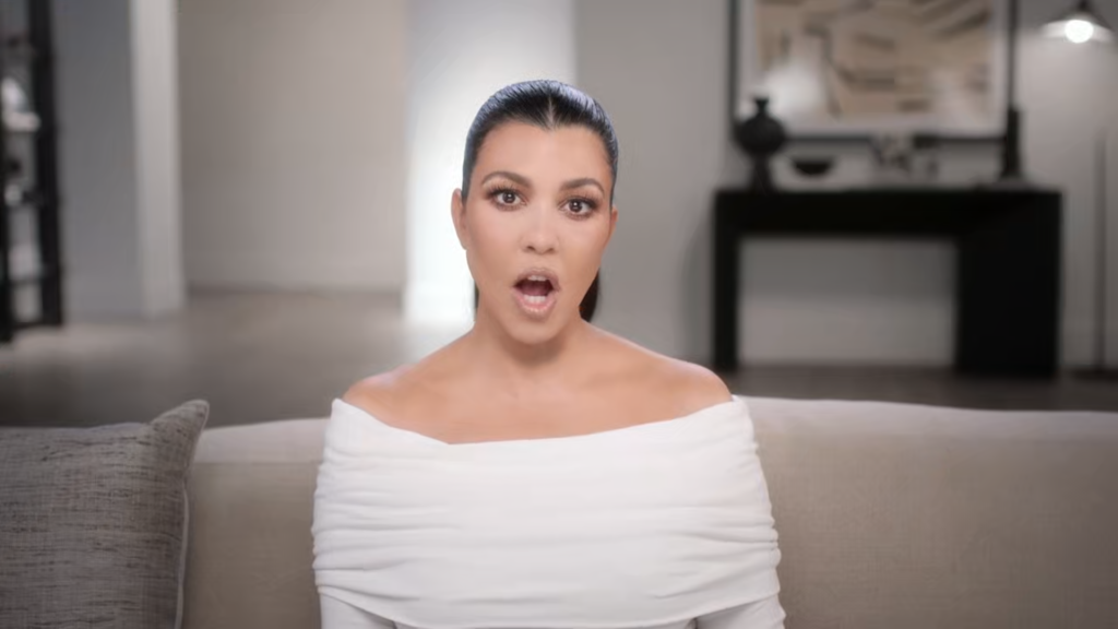 Kourtney Kardashian tells the confessional camera what she thinks with her mouth open, wearing a broad -necked white blouse.