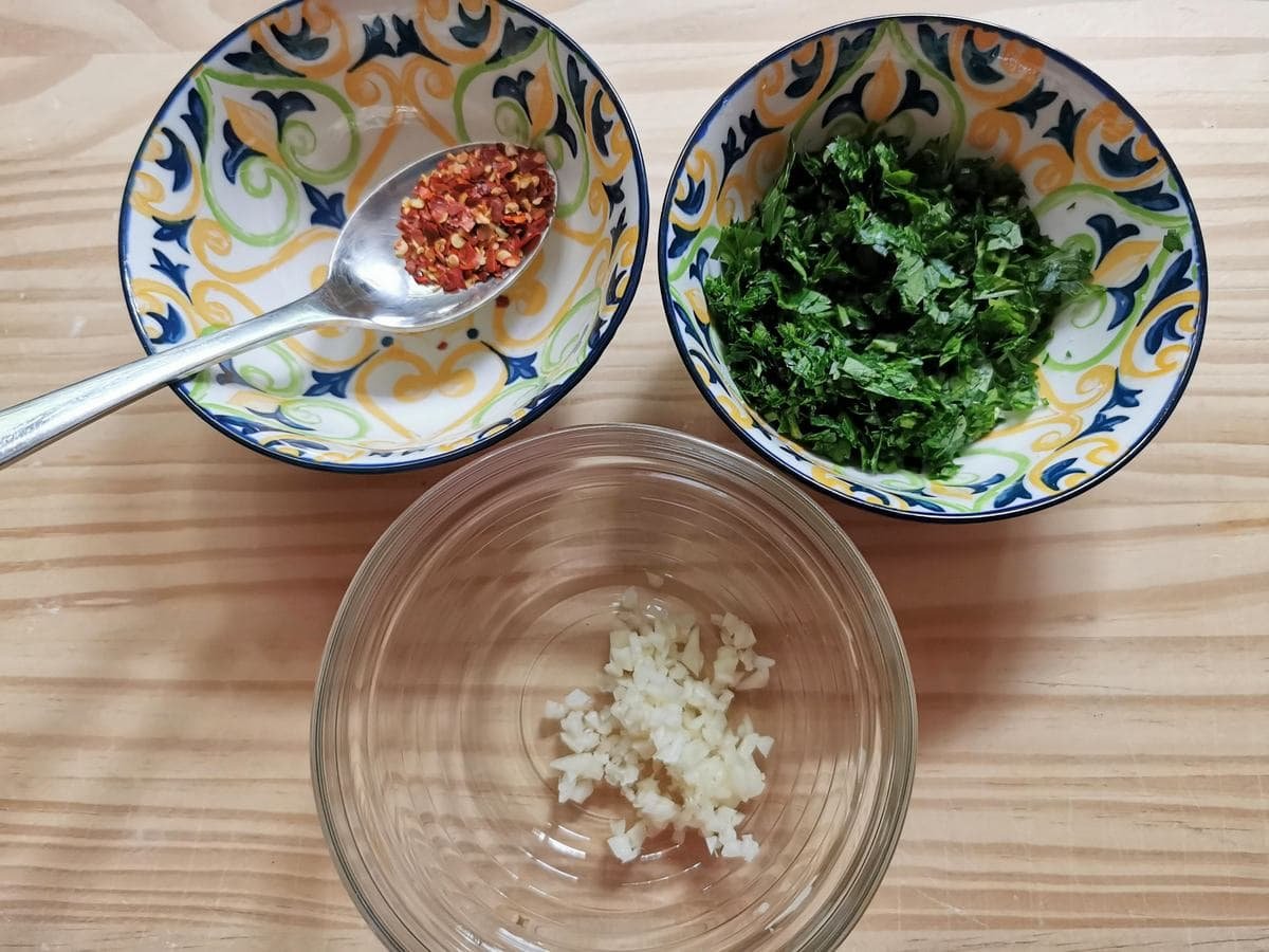 Chopped fresh garlic in a bowl, chopped fresh parsley in a small bowl and peperoncino flakes in a third small bowl.