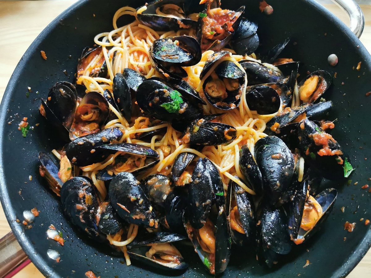 Cooked spaghetti mixed with cooked clams and tomato sauce in large frying pan.